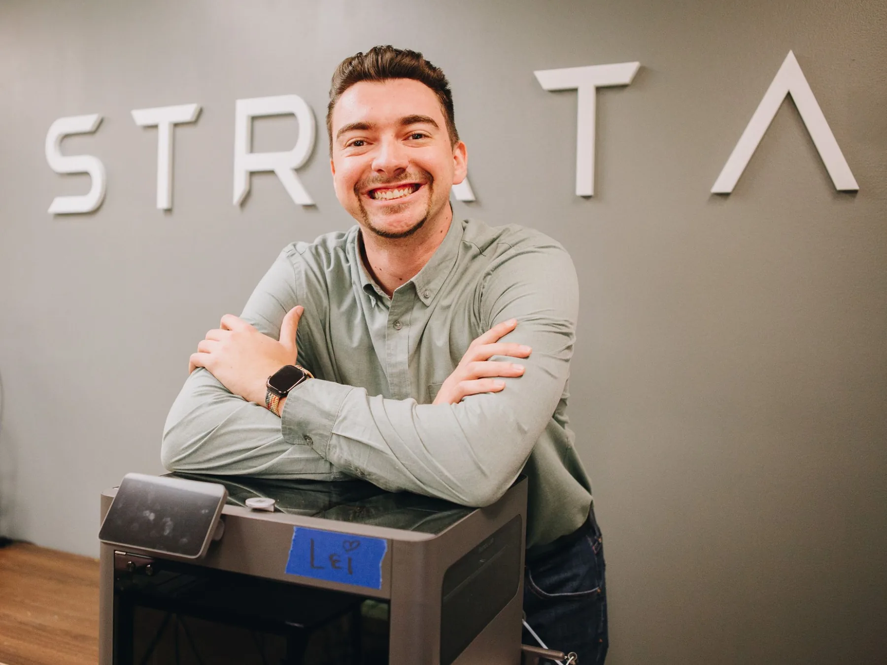 Benjamin, founder of Strata Printing, smiling and leaning on a 3D printer in front of the Strata sign